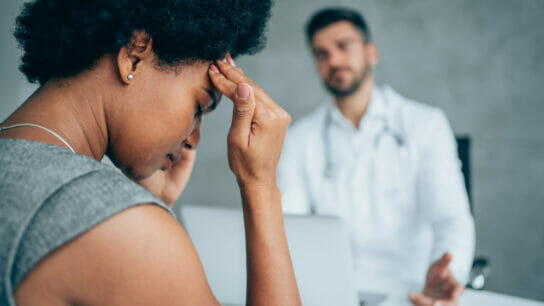 African-american_woman_sitting_opposite_the_doctor_in_ his_office_and_ holding_her_head_with_ hands.