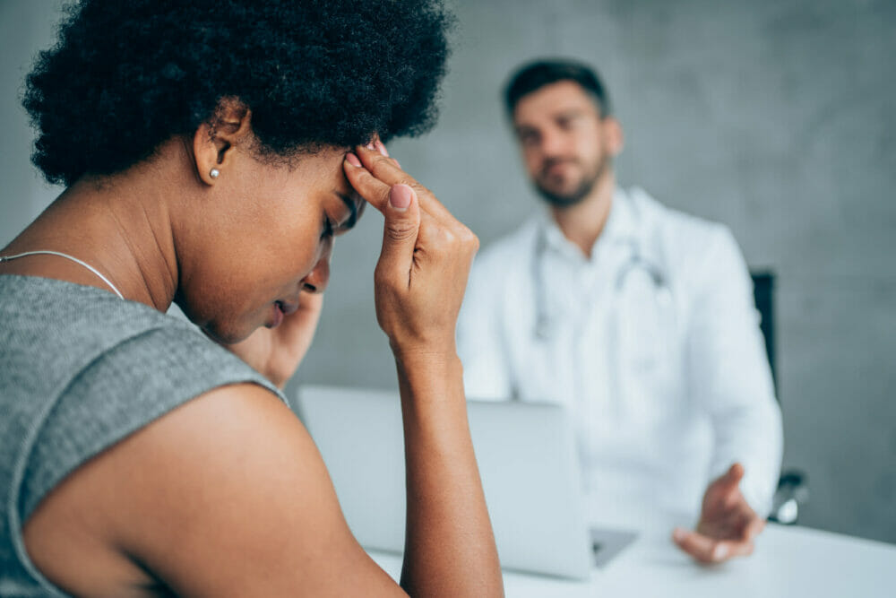 African-american_woman_sitting_opposite_the_doctor_in_ his_office_and_ holding_her_head_with_ hands.
