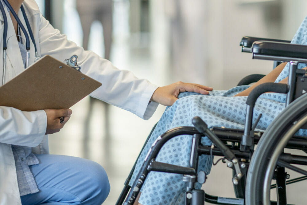 Individual_with_disability_sits_in_wheelchair_as_doctor_approaches_with_hand_on_their_knee_in_empathetic_manner.