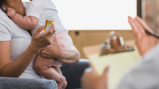 An-unrecognizable-new-mom-with-her-baby-holds-the-pill-bottle-as-she-discusses-the-side-effects-with-an-unrecognizable-male-psychiatrist.