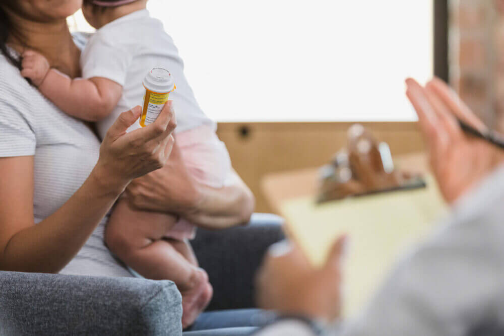 An-unrecognizable-new-mom-with-her-baby-holds-the-pill-bottle-as-she-discusses-the-side-effects-with-an-unrecognizable-male-psychiatrist.