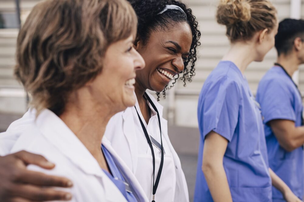 Happy-female-physician-with-arm-around-colleague-outside-hospital
