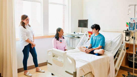 Wide-shot-of-senior-female-doctor-consulting-with-teenage-boy-during-visit-to-hospital-emergency-room.